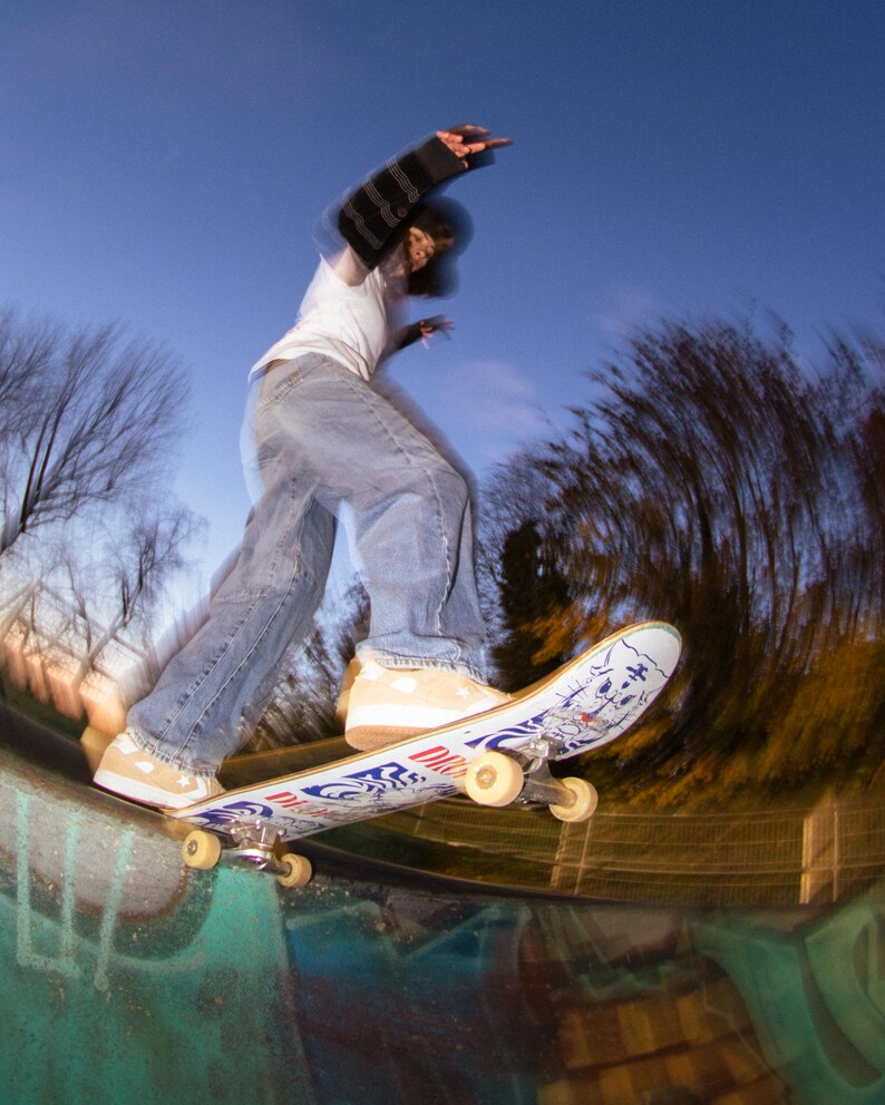 May include: A skateboarder in mid-air, performing a trick at a skate park. The skateboard has a white deck with blue and red graphics. The skater wears a white t-shirt, blue jeans, and tan sneakers. The background is a blurred view of trees and a blue sky.