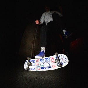May include: A skateboard with a white deck featuring blue and red graphics and the words "DROP IN". A person is in mid-air, wearing blue sneakers, black pants, and a light-colored long-sleeve shirt. The scene is dark.