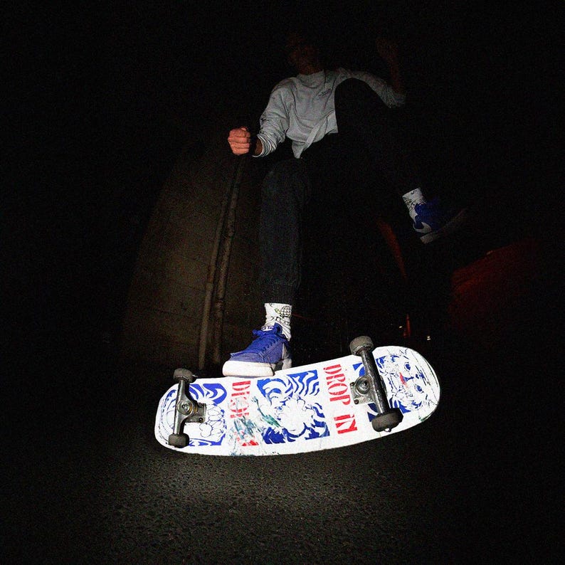 May include: A skateboard with a white deck featuring blue and red graphics and the words "DROP IN". A person is in mid-air, wearing blue sneakers, black pants, and a light-colored long-sleeve shirt. The scene is dark.