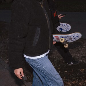May include: A black, puffy, zip-up jacket with a high collar. The person is wearing light blue jeans and a white t-shirt. A skateboard with a blue and white design is visible.