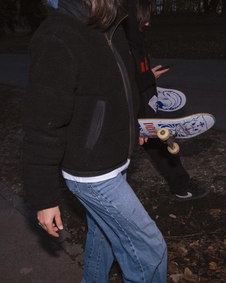 May include: A black, puffy, zip-up jacket with a high collar. The person is wearing light blue jeans and a white t-shirt. A skateboard with a blue and white design is visible.