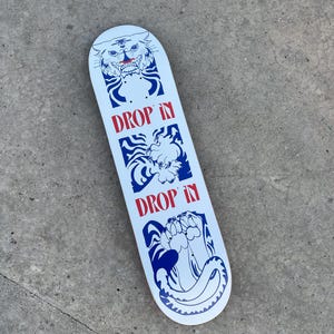 May include: White skateboard with a blue and red graphic design. The design includes a tiger, a creature, and a paw with the words "DROP IN" in red. The skateboard is on a gray surface.