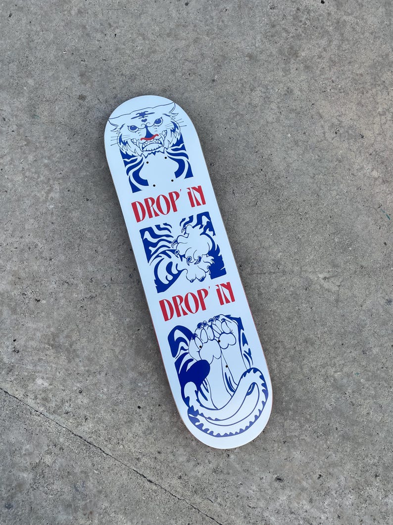 May include: White skateboard with a blue and red graphic design. The design includes a tiger, a creature, and a paw with the words "DROP IN" in red. The skateboard is on a gray surface.
