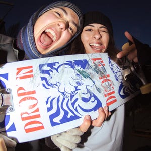 May include: A white skateboard with blue and red graphics, including the words "DROP IN" and a tiger illustration. The skateboard is held by two people, one wearing a beanie and the other a scarf.