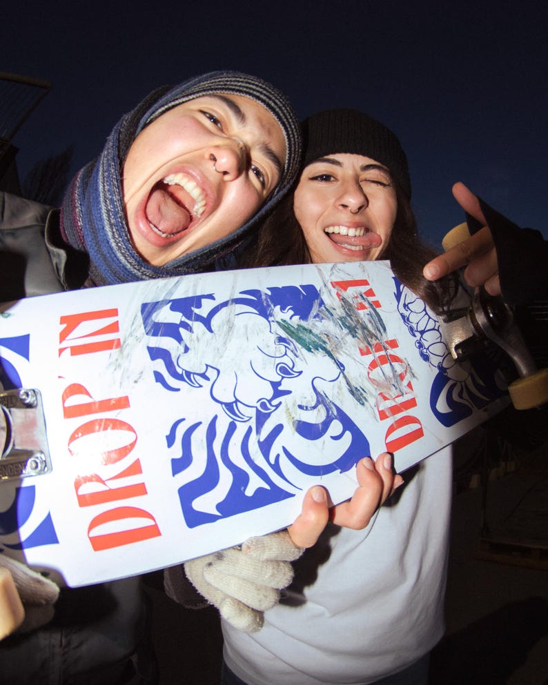May include: A white skateboard with blue and red graphics, including the words "DROP IN" and a tiger illustration. The skateboard is held by two people, one wearing a beanie and the other a scarf.