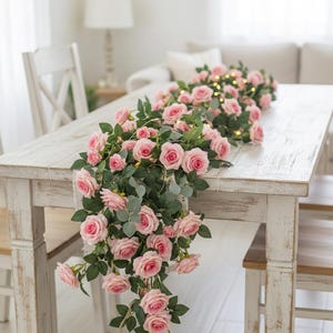 Peut inclure: Une table en bois blanc vieilli est ornée d'une guirlande de roses roses et de feuilles vertes. Les roses sont à différents stades de floraison, avec de petites lumières de fées entrelacées. Deux chaises blanches sont visibles.