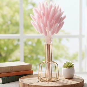 Peut inclure: Un arrangement décoratif avec un vase en fil d'or contenant un bouquet d'herbe de queue de lapin rose. Le vase est posé sur un plateau rond en bois, à côté d'une petite plante succulente en pot et d'une pile de livres.