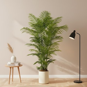 Peut inclure: Un grand palmier artificiel vert dans un pot blanc. À droite, un lampadaire noir avec une lumière blanche. À gauche, une petite table d'appoint en bois avec un vase blanc et des livres.