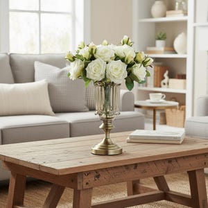 May include: A display of white roses in a clear, decorative glass vase with a brass base. The vase is placed on a rustic wooden coffee table. The roses have green leaves. The table is in a living room.