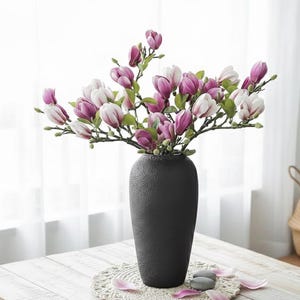 Op de afbeelding: Een donkergrijze vaas met takken van roze en witte magnoliabloemen. De vaas staat op een witte houten tafel, met een decoratief kleedje en verspreide bloemblaadjes. De bloemen hebben groene bladeren.