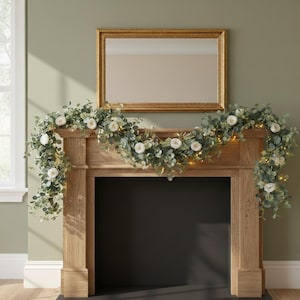 Peut inclure: Un manteau de cheminée en bois avec un miroir encadré d'or au-dessus. Une guirlande de fleurs blanches, de feuilles vertes et de petites lumières orne le manteau. L'ouverture de la cheminée est noire.
