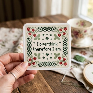 May include: A square cross-stitch design with the text "I overthink therefore I am." The design features a cream background with a green border, red berries, green leaves, and small green hearts and stars. The design is held by a person.