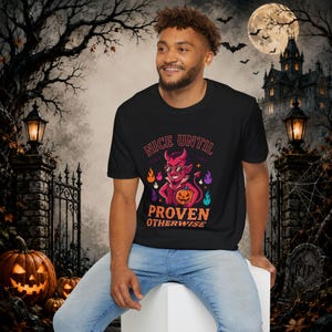 May include: A black short-sleeve t-shirt featuring a graphic of a red devil wearing sunglasses and holding a pumpkin. The text reads "NICE UNTIL PROVEN OTHERWISE" in orange and red letters, surrounded by small colorful flames and bat silhouettes.