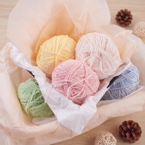 May include: Five balls of soft, plush yarn in pastel shades of yellow, light pink, bubblegum pink, mint green, and baby blue, nestled inside a container lined with light peach-colored tissue paper, accompanied by two small brown pinecones.