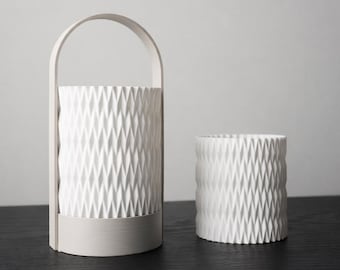 May include: Two white ceramic vases with a geometric diamond pattern. One vase has a handle and a rectangular base. The other is a smaller, cylindrical vase. Both are on a dark surface.