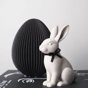 Puede incluir: Una figura de conejo de cerámica blanca con una pajarita negra se encuentra junto a un gran objeto decorativo negro en forma de huevo. El huevo tiene una textura acanalada. Los objetos están sobre una superficie negra.