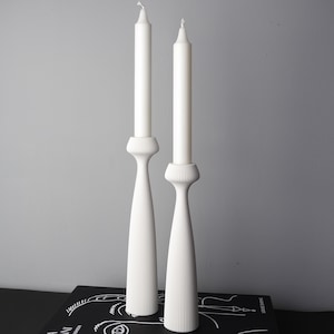 Könnte beinhalten: Zwei weiße Kerzenhalter mit weißen Kerzen. Die Kerzenhalter haben ein geripptes Design am Sockel und eine ausgestellte, längliche Form. Die Kerzen sind hoch und schlank. Der Hintergrund ist einfarbig grau.