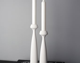 May include: Two white candle holders with white candles. The candle holders have a ribbed design on the base and a flared, elongated shape. The candles are tall and slender, with a slightly curved top. The background is a solid gray.