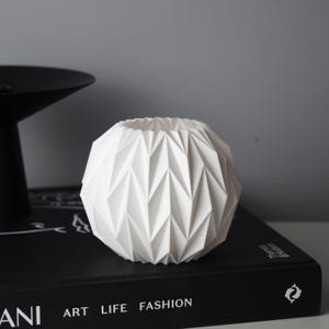 Könnte beinhalten: Eine weiße, geometrische, kugelförmige Vase mit einem strukturierten, eckigen Design. Die Vase steht auf einem schwarzen Buch mit den Worten "ART LIFE FASHION". Im Hintergrund befindet sich ein schwarzes, rundes Objekt.