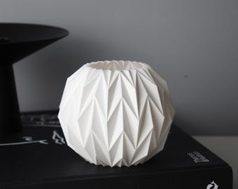 May include: A white, geometric, spherical vase with a textured, angular design. The vase is sitting on a black book with the words "ART LIFE FASHION" visible. A black, round object is in the background.