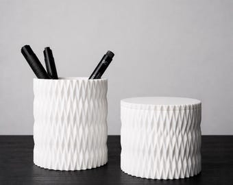 May include: Two white ceramic containers with a geometric diamond pattern. One cylindrical container holds three black pens. The other is a smaller, lidded container. Both are on a dark surface, against a light gray background.