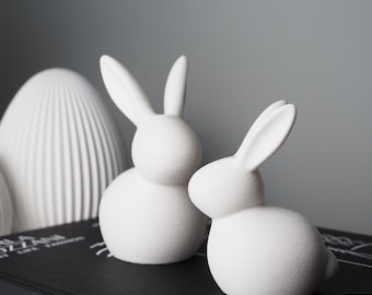 May include: Two white ceramic bunny figurines with long ears and rounded bodies. The bunnies are placed on a black surface, with a white, textured, egg-shaped decorative object in the background. The bunnies are a decorative item.