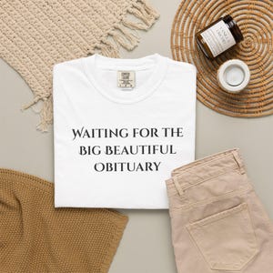 May include: A white short-sleeved t-shirt with a crew neckline featuring the black printed text "WAITING FOR THE BIG BEAUTIFUL OBITUARY" centred on the chest. The garment has a Comfort Colors brand label visible at the inner neck area.