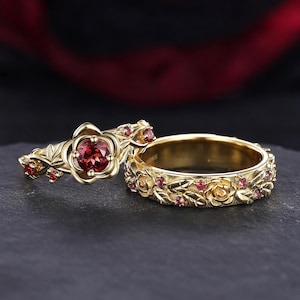 Puede incluir: Un conjunto de anillos de oro que presenta una piedra preciosa roja redonda central rodeada de pétalos y hojas de rosa dorados esculpidos, junto a una banda de oro a juego con un patrón continuo de pequeñas rosas y piedras rojas incrustadas.