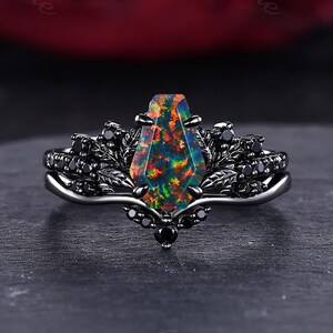 May include: A black metal ring featuring a central coffin-shaped opal gemstone with vibrant orange, green, and red flashes. The band is detailed with intricate leaf motifs and small, round black stones set along the sides and the base of the setting.