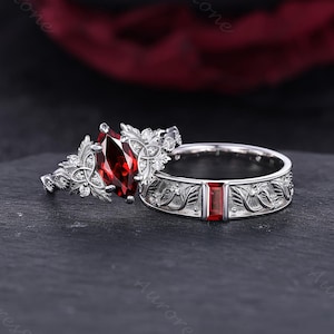 May include: A silver ring set featuring a marquise-cut deep red gemstone flanked by intricate leaf-like metalwork and small clear stones, paired with a matching band containing a single rectangular red stone and engraved floral patterns.