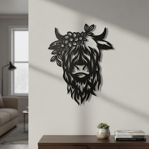 May include: Black metal wall art of a Highland cow head, adorned with a floral crown. The cow's shaggy fur and facial features are intricately cut out. The artwork is mounted on a white wall, adding a rustic touch to the decor.