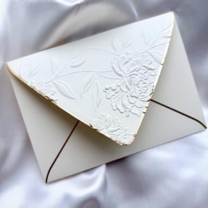 May include: A white envelope with a floral embossed design and gold trim. The envelope is set against a white satin background, creating a luxurious and elegant aesthetic. The design features a detailed floral pattern.