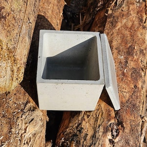 Könnte beinhalten: Eine quadratische, hellgraue Betonbox mit passendem Deckel, teilweise geöffnet. Die Box steht vor einem Hintergrund aus rauen, braunen Holzstämmen. Die Box hat ein schlichtes, modernes Design.