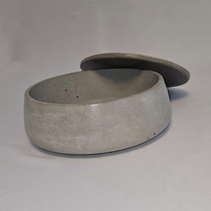 Könnte beinhalten: Eine hellgraue Betonschale mit passendem Deckel. Die Schale ist oval geformt mit abgerundetem Boden und glatter, matter Oberfläche. Der Deckel ist eine flache, kreisförmige Scheibe, etwas dunkler gefärbt.