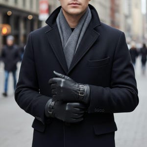Puede incluir: Una persona con un abrigo azul marino, una bufanda gris y guantes de cuero negros. Los guantes tienen una hebilla. El abrigo tiene un diseño clásico con solapa de muesca y cierre de un solo botón. El fondo es una calle borrosa.