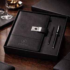 May include: A black leather journal and pen set in a black gift box. The journal has a silver clasp and the word "ORAVIA" printed on the front. A black pen and a thin black pen are included.
