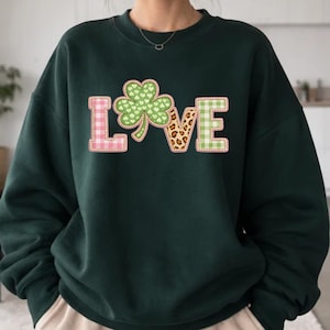 May include: Dark green sweatshirt featuring the word "LOVE" in a decorative design. The "L" is pink gingham, the "O" is a green shamrock, the "V" is leopard print, and the "E" is green gingham. A St. Patrick's Day themed top.