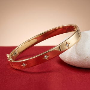 May include: A gold-colored bangle bracelet with star-shaped accents. The bracelet is displayed on a red surface with a white stone in the background. The bracelet has a clasp closure and is a classic jewelry piece.