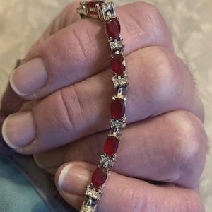 May include: A silver-toned tennis bracelet featuring a series of oval-cut deep red gemstones alternating with small clusters of clear white stones. The bracelet has a flexible link construction and a metallic finish throughout its length.