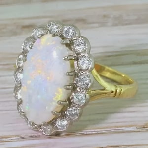 May include: An oval-shaped opal ring featuring a central iridescent white opal stone surrounded by a halo of small round clear diamonds. The setting is crafted from yellow gold, with the diamonds mounted in a white metal frame for contrast.