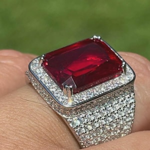 Könnte beinhalten: Ein silberfarbener Ring mit einem großen, rechteckigen, tiefroten Mittelstein, der von vier Krappen gehalten wird. Die rechteckige Fassung und die breite Ringschiene sind mit zahlreichen kleinen, klaren, funkelnden Steinen besetzt.