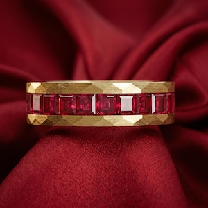May include: A gold band ring featuring a central channel set with a row of square-cut red rubies. The gold edges of the ring have a textured, faceted finish, creating a geometric pattern that frames the vibrant red gemstones in a horizontal line.