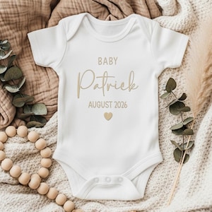 May include: A white short-sleeved baby bodysuit featuring the text BABY Patrick AUGUST 2026 in a tan script and sans-serif font, centred above a small tan heart icon. The garment has a round neckline and three snaps at the bottom closure.