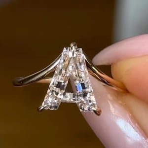 May include: A gold ring featuring a prominent letter A centerpiece crafted from multiple clear, faceted stones. The letter A is set atop a thin, smooth gold band that curves slightly upward to meet the base of the letter, creating a distinct silhouette.
