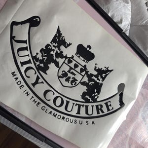May include: A white bag with the words "Juicy Couture" in a black, stylised font. The bag features a black emblem with a crown and shield design, and the text "Made in the Glamorous USA".