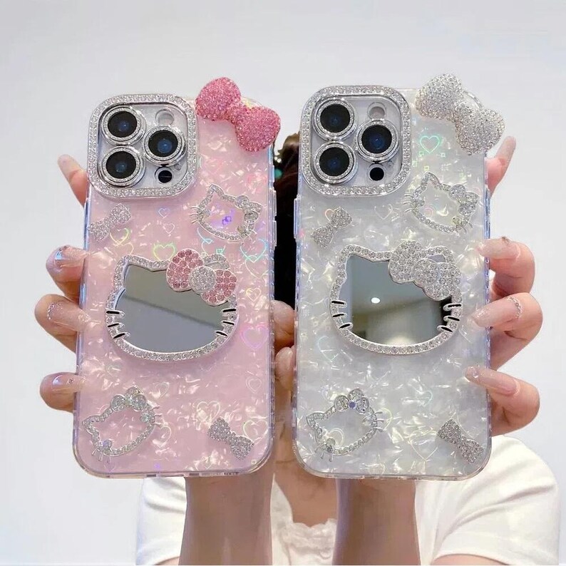 May include: Two phone cases, one pink and one white, each adorned with sparkling embellishments. The cases feature a Hello Kitty design with a mirror, bows, and rhinestone accents. The camera lens is surrounded by a ring of rhinestones.