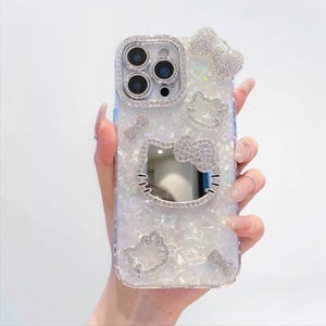 May include: A clear phone case with a pearlescent finish, adorned with sparkling rhinestones. The case features a Hello Kitty design, bows, and a decorative frame around the camera lenses. The case is held in a hand.