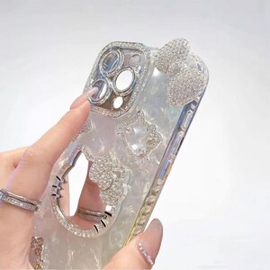 May include: A clear phone case with a pearlescent finish, adorned with sparkling rhinestones. The case features a bow, a Hello Kitty design, and a camera lens frame, all embellished with crystals. The case is designed to fit a smartphone.