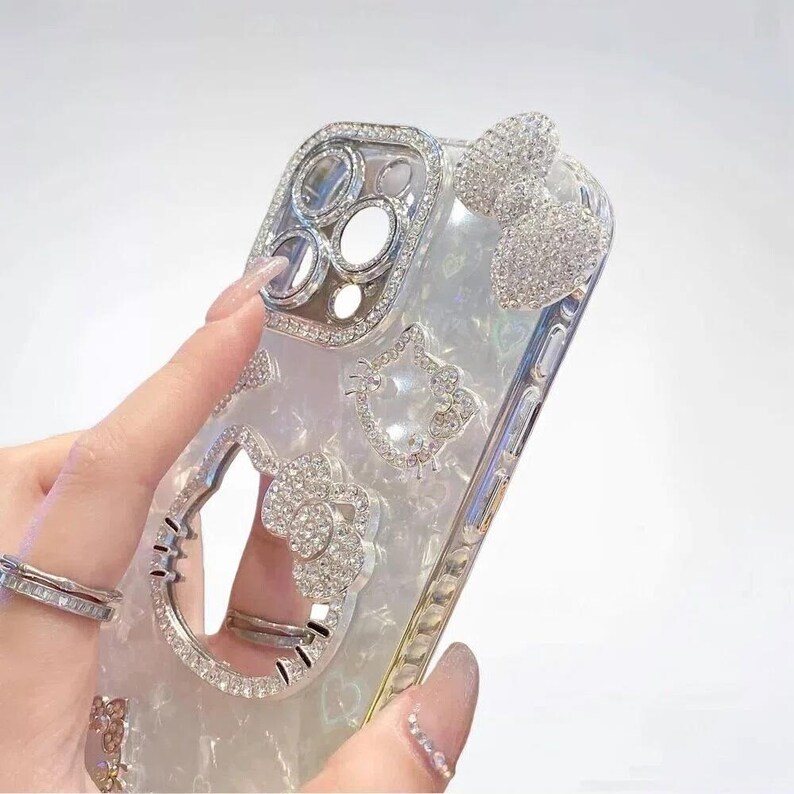 May include: A clear phone case with a pearlescent finish, adorned with sparkling rhinestones. The case features a bow, a Hello Kitty design, and a camera lens frame, all embellished with crystals. The case is designed to fit a smartphone.