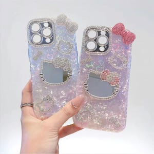 May include: Two iridescent phone cases with Hello Kitty designs. Each case features a mirrored Hello Kitty face, sparkling bows, and rhinestone accents. The cases have a pearlescent finish with heart and bow patterns.